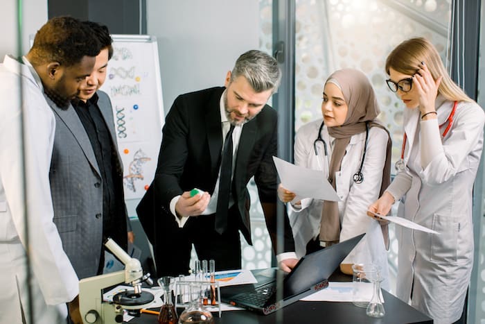 Mature man in suit, pharmaceutical company director, holding a bottle with new medicine drug, smiling, while talking with multiethnic team of doctors researchers, about clinical trial.