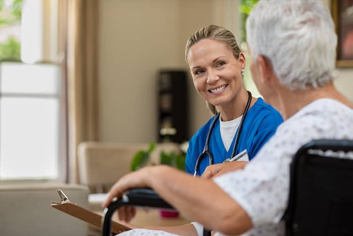 Friendly doctor examining health of patient sitting in wheelchair. Happy smiling nurse consulting disabled patient about treatment. Nurse caring about elder handicap man at home.