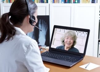 Female doctor in her surgery office with headset in front of her laptop, an x-ray of a foot in hand, talking with a senior patient, telemedicine concept