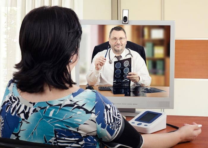 Middle-aged cardiologist sits face to face with online female patient which measures her blood pressure. Handsome virtual doctor attentively reviews brain x-ray results with her. Horizontal shot on blurry indoors background
