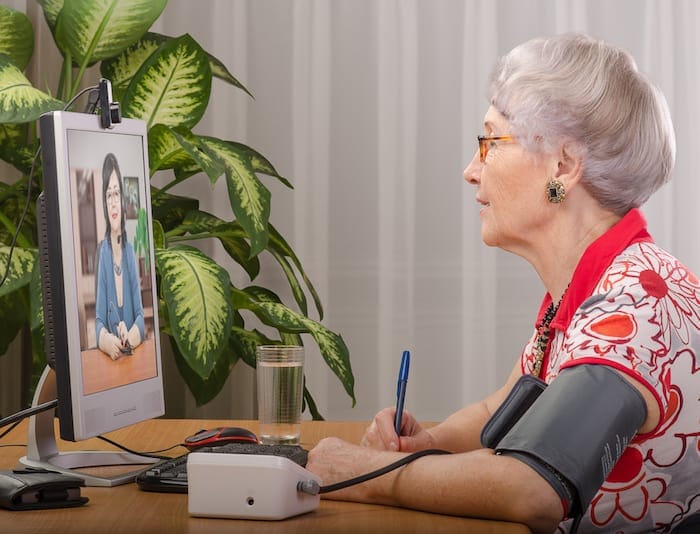 Old grey haired wom an is measuring blood pressure by herself during virtual doctor visit. Glasses senior woman sitting opposite monitor. On the screen, telehealth doc is consulting her. Side shot