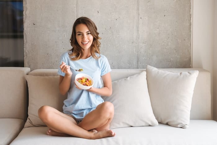 Delighted young woman eating healthy breakfast while sitting on a couch at home