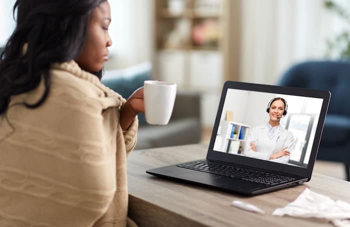 healthcare, technology and people concept - african american sick woman in blanket having video call with doctor on laptop computer and drinking tea at home