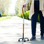 Senior disabled caucasian woman hands on cane outside nursing home park. Close up of elderly lady holding a walking stick outdoors of healthcare facility on the sunny day.