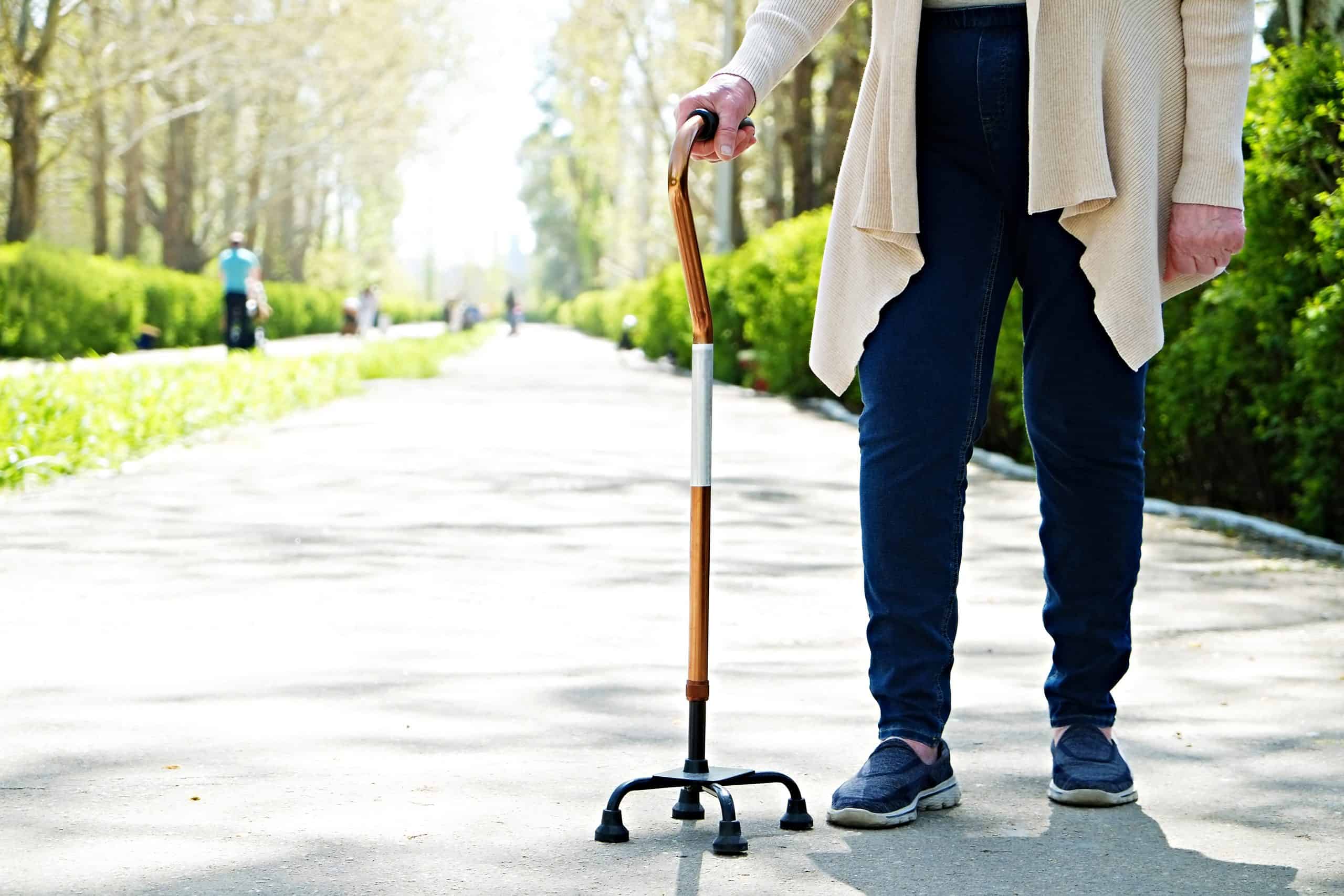 Senior disabled caucasian woman hands on cane outside nursing home park. Close up of elderly lady holding a walking stick outdoors of healthcare facility on the sunny day.