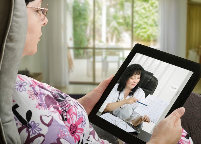 Mature adult woman consults a telemedicine doctor with tablet computer sitting in soft chair. In touchscreen, female doctor in white uniform reviewing blood pressure report. With telehealth application patient can reach relevant specialist remotely. Horizontal side shot on indoors blurred background