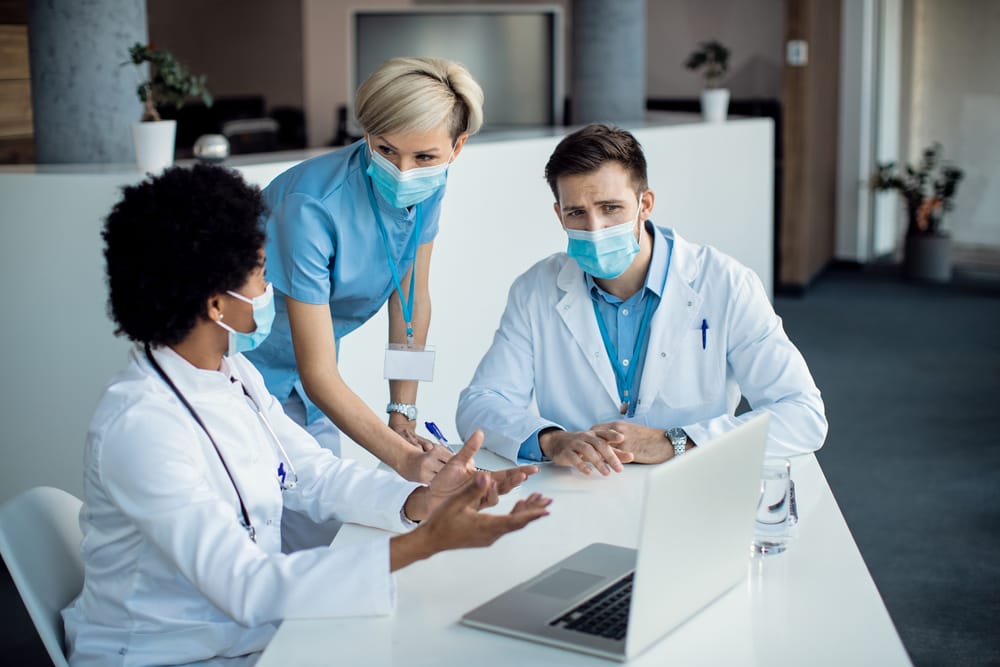 Team of healthcare workers cooperating while using laptop on a meeting at medical clinic. They are wearing face masks due to coronavirus pandemic.