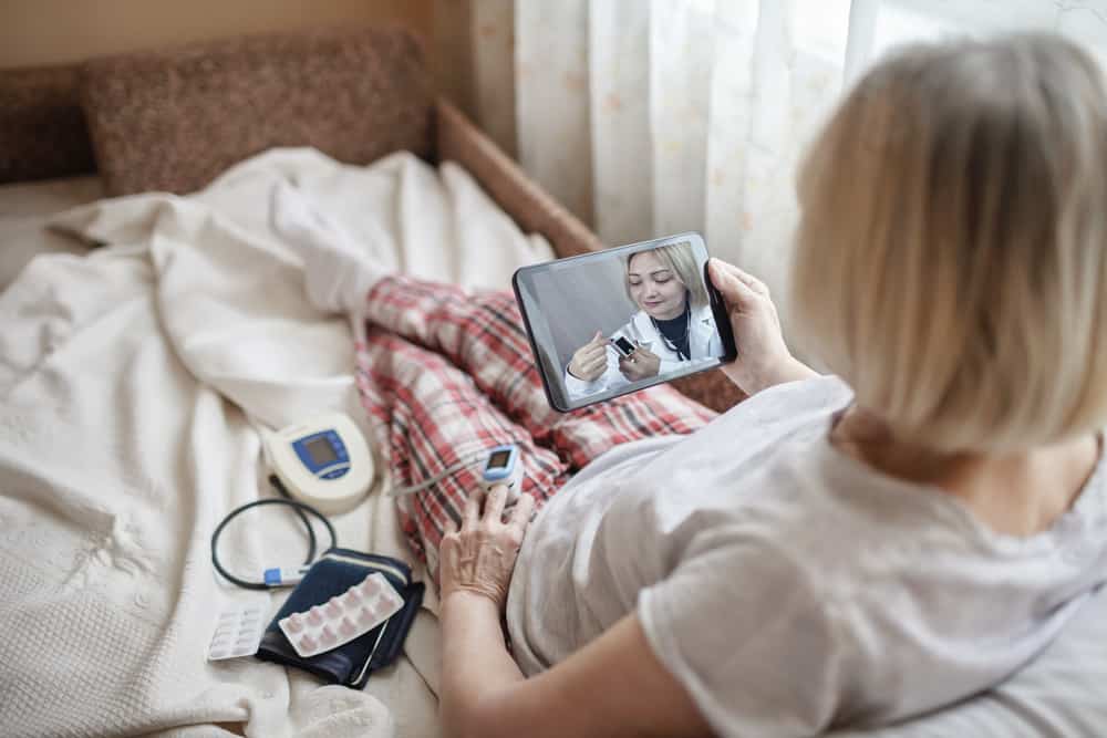 Old woman in bed looking at screen of laptop and consulting with a doctor online at home, telehealth services during lockdown, distant video call, modern tech healthcare application