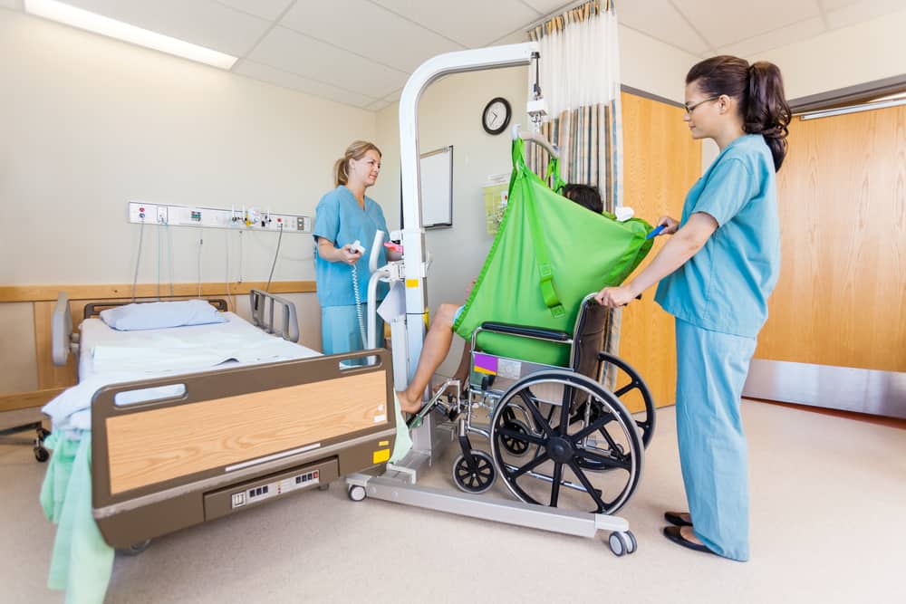 Female nurses transferring male patient from hydraulic lift to wheelchair in hospital