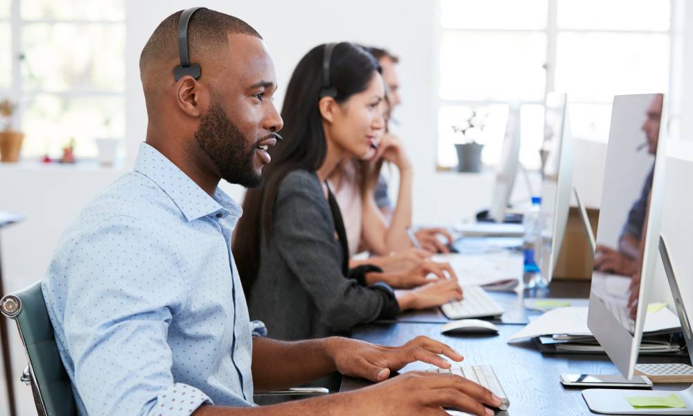 A team of three IT professionals sitting at a long desk in an office while working on computers and using headsets.