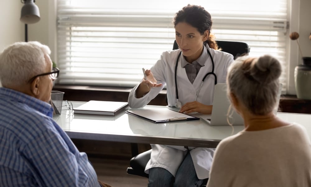 A doctor sits behind a desk with a clipboard and a laptop as she communicates with an older couple during an appointment.