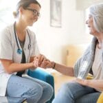 A doctor wearing a stethoscope around her neck talks to an older woman with a gray sweater around her shoulders.