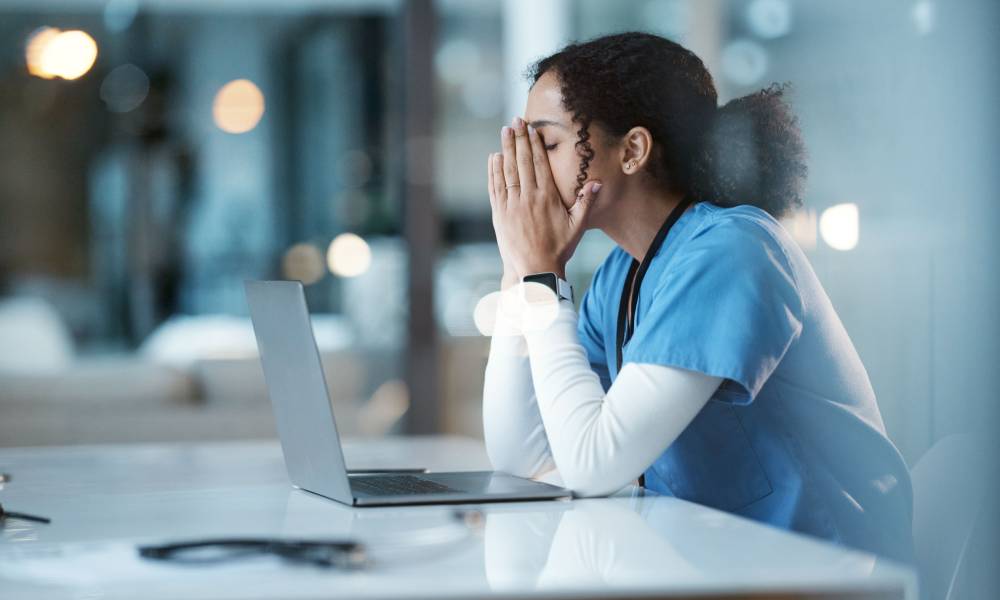 A young healthcare professional in scrubs leaning on her desk and holding her face in her hands due to stress.