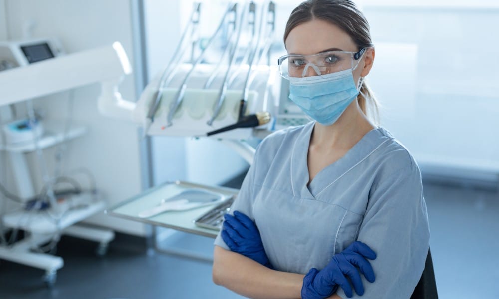 A young female dentist wearing a mask, eyewear, latex gloves, and scrubs standing with her arms crossed in a dental exam room.