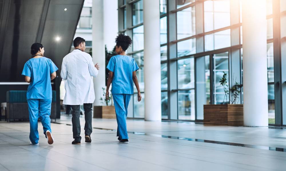 The backside of two doctors wearing blue scrubs and one wearing a white coat walking down a hospital hallway with windows.