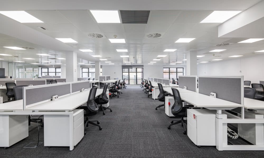 Connected white desks form rows in an office space. Gray partitions divide the desks, and black chairs appear at the desk.
