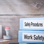 Two blue binders labeled "Safety Procedures" and "Work Safety" sitting on a desk next to a mug and papers.