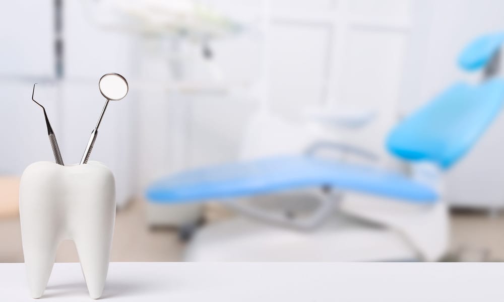 A close-up view shows two dental instruments sticking out the top of a fake tooth. A dental office is blurry in the back.