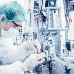 Medical worker wearing safety gear and a hairnet touching a large medical device inside a machinery room.