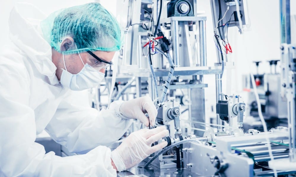 Medical worker wearing safety gear and a hairnet touching a large medical device inside a machinery room.