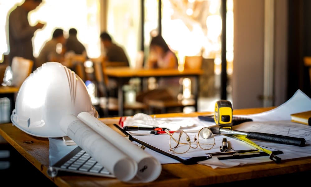 A wooden table contains a white hard hat, glasses, a tape measure, rolled-up paper, a hammer, blueprints, and a calculator.