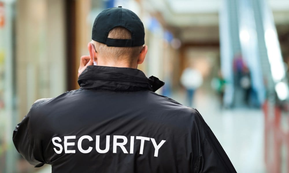 A security guard in a black jacket and hat walking the halls of a medical facility. The escalator is in front of him.
