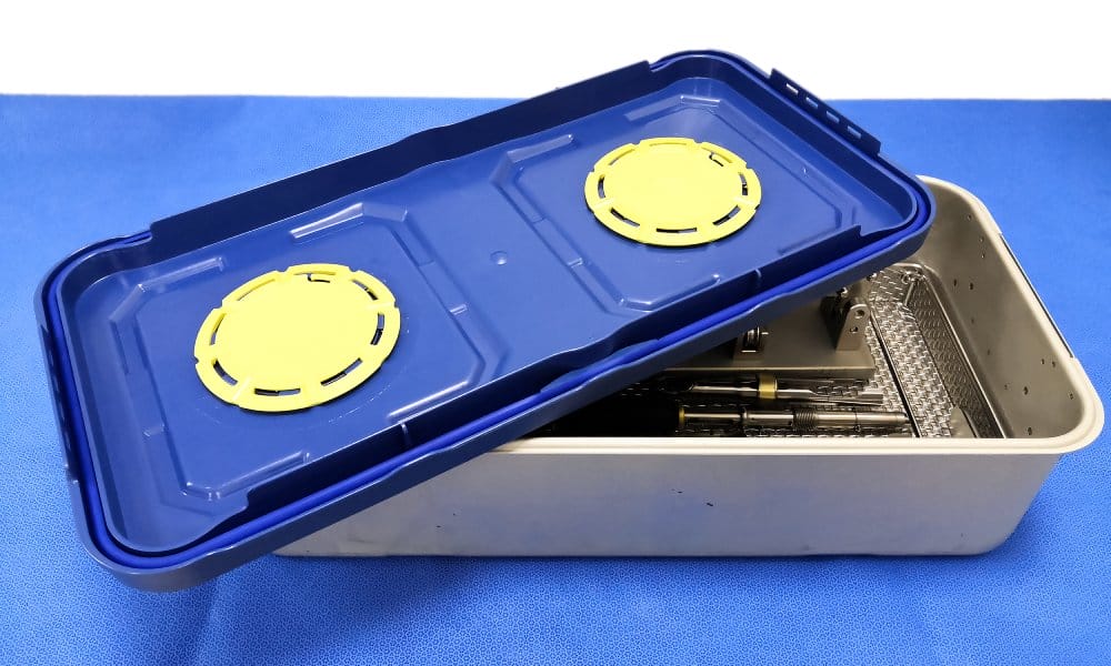 An aluminum sterilization case contains a steel basket with medical instruments. A blue lid with yellow circles sits on top.
