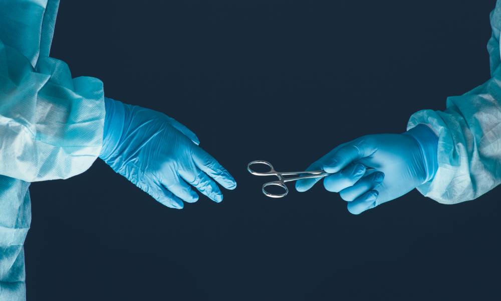 A close-up view shows a healthcare professional dressed in surgical garments passing surgical scissors to another professional.