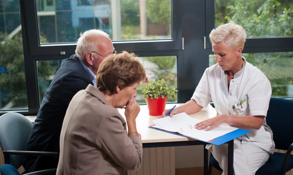 A nurse in a white outfit sits at a table across from an elderly couple. She reviews the paperwork in a blue notebook.
