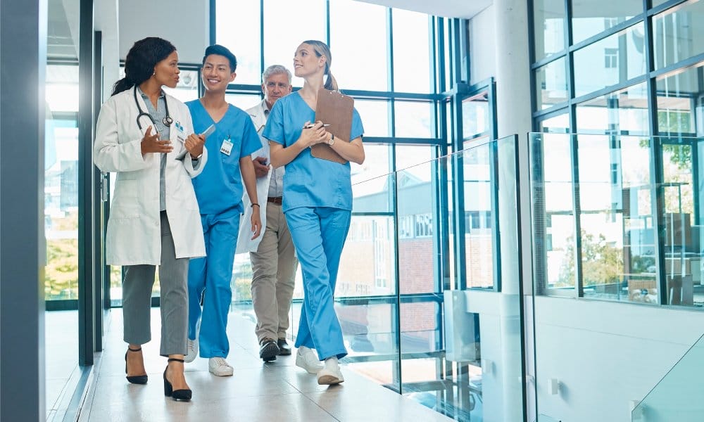 Four adults talking together while walking down a hall surrounded by windows. Two are wearing scrubs and two have white coats.