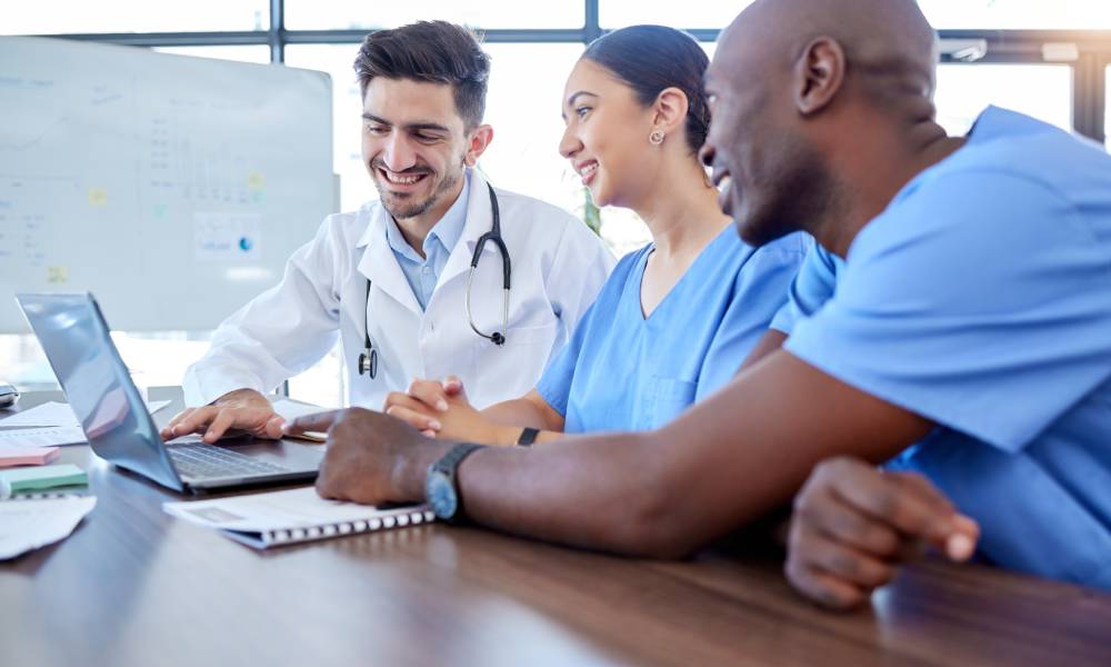 Three healthcare professionals—one in a white coat and two in scrubs—sitting around a laptop and looking at the screen.