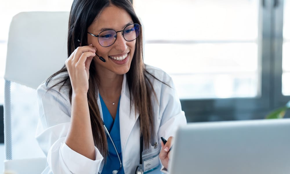 A female healthcare professional sits at a desk, smiling at an open laptop. She is touching an earpiece in her ear.