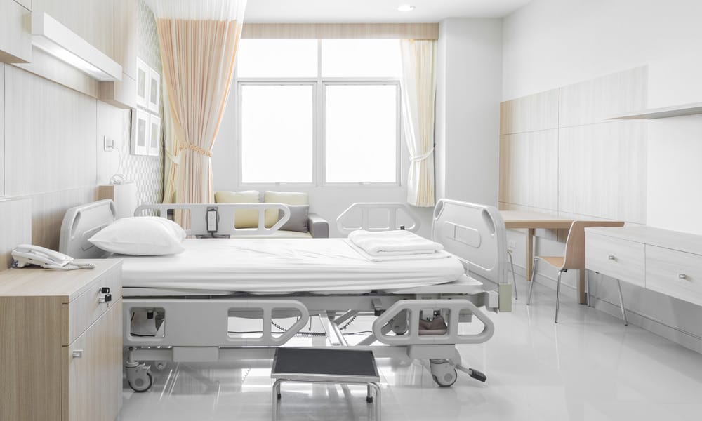 Bright hospital room with white linen on the bed, brown privacy curtains, a table, and a large window in the back.