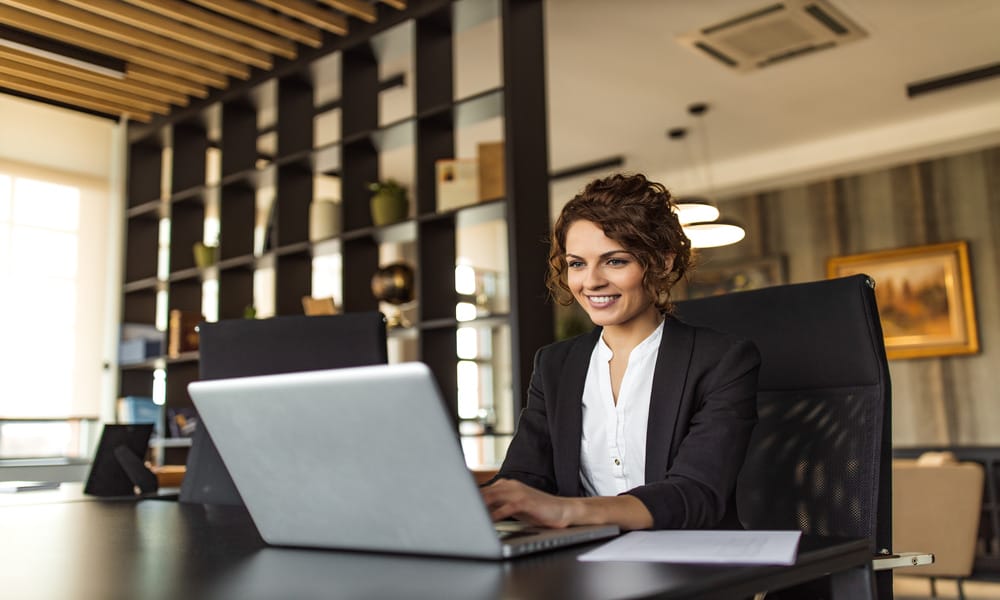 A smiling woman in a professional business suit sits at a desk in a black chair, typing on a silver laptop.