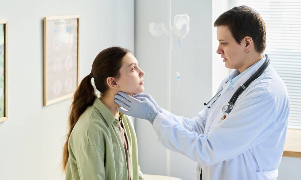 A person wearing latex gloves and a stethoscope touches either side of another person's throat in a healthcare facility.