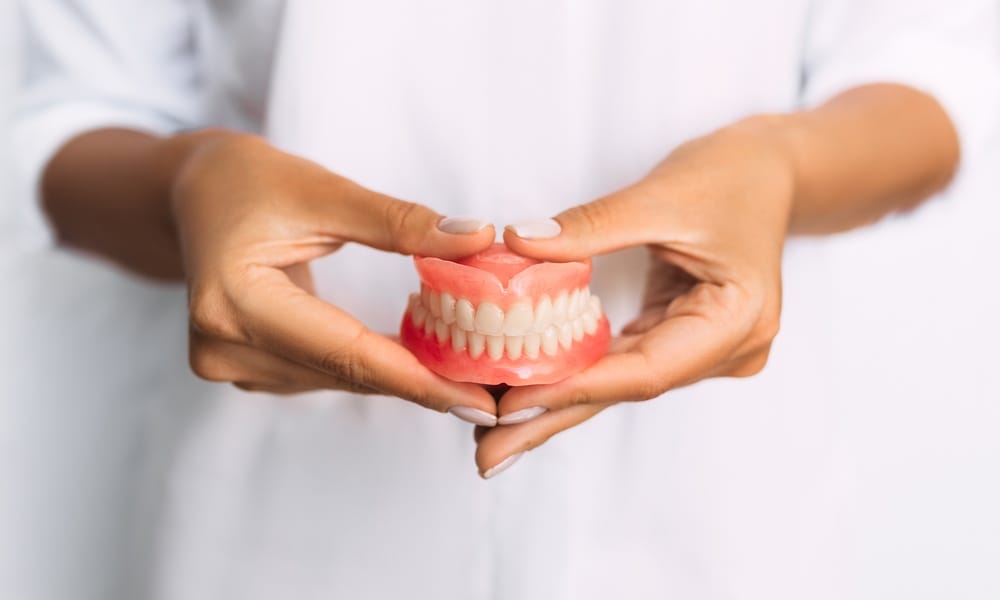 A close-up view shows a person in a white lab coat holding a set of dentures in their hands. They have tan fingernail polish.