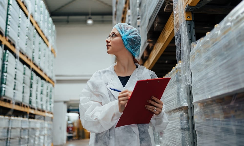 A woman wearing a disposable lab jacket and hair net holds a clipboard and walks down a medical warehouse aisle observantly.