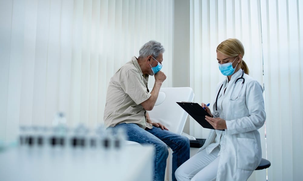 A man in a disposable mask sitting on a hospital bed and turning his head to cough while a doctor takes notes.