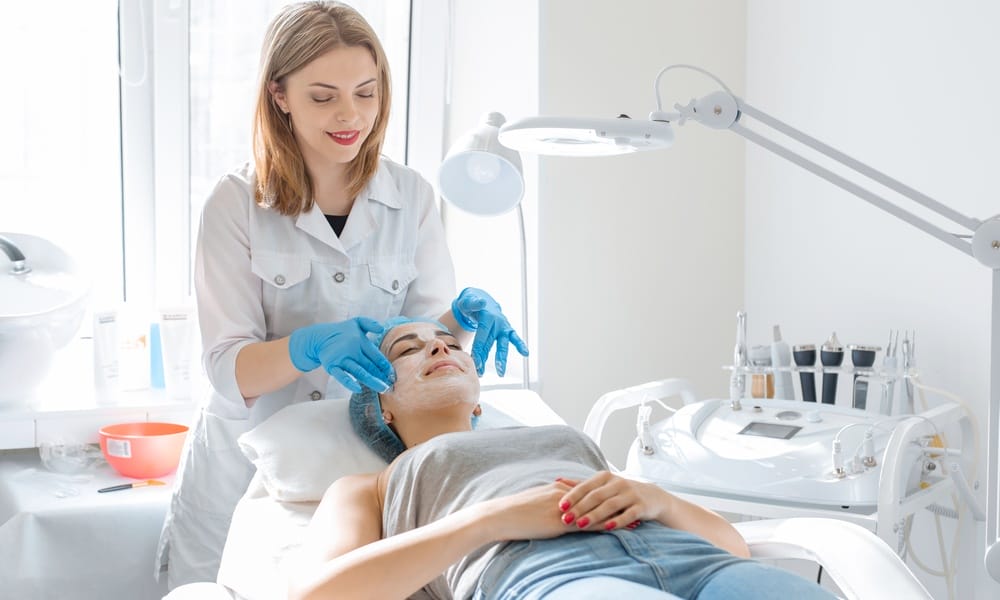 A professional wearing a white coat and blue gloves working a facial product into the skin of a client laying on a medical bed.