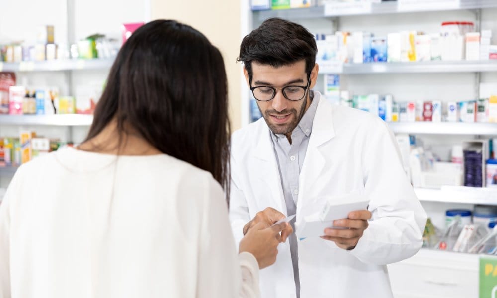 A pharmacist gesturing to a card and explaining a prescription to a patient picking up their medication.