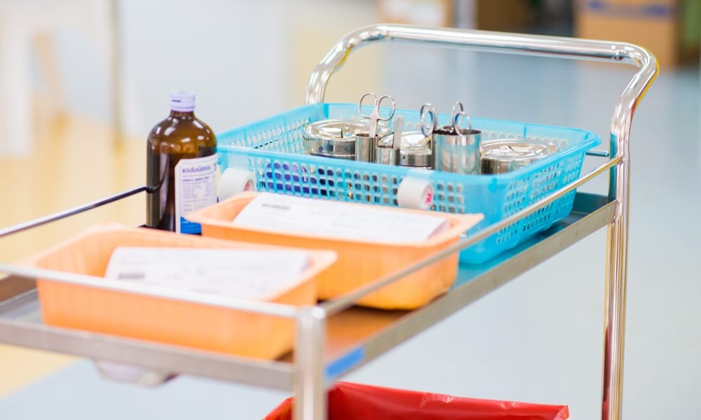 A multitiered medical cart sitting in a hospital hallway. On the top rack, there are baskets filled with medical supplies.