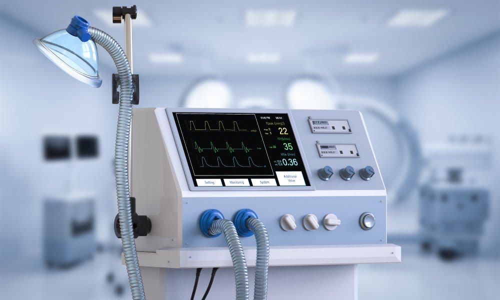 A close-up of a medical ventilator machine in a hospital. The black screen displays vitals information.