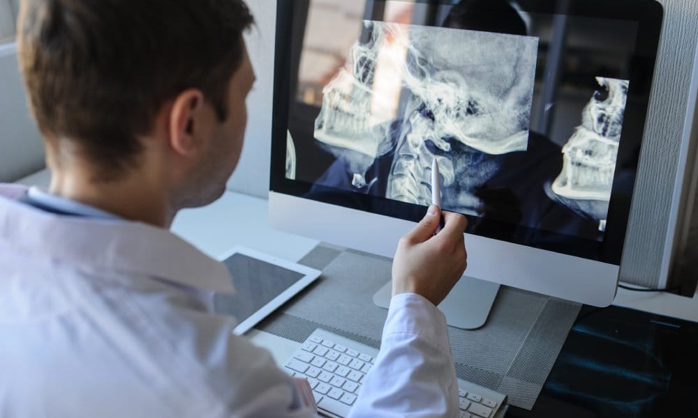 A male radiologist examines neck X-rays on a computer and points with a pen to the cervical vertebrae.