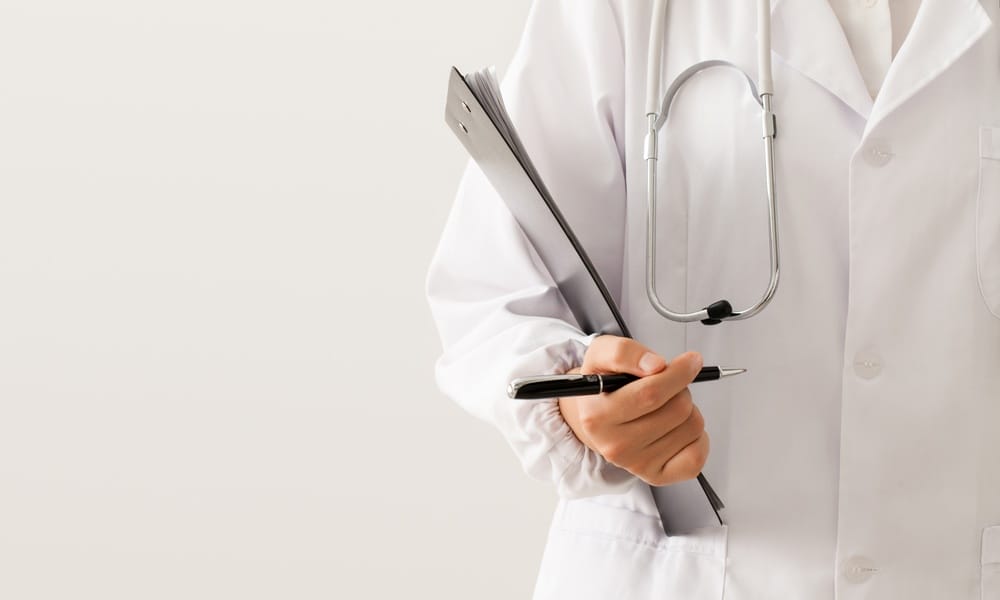 A close-up of a person in a white lab coat and a stethoscope, holding a pen and a clipboard.