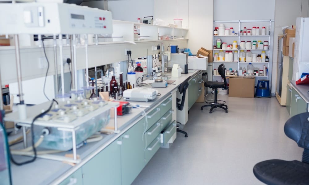 A laboratory workspace containing shelves of bottles, testing equipment, lab chairs, and organized workstations.