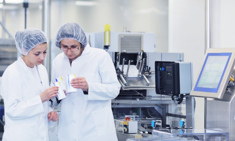 Two medical professionals in lab coats and hair coverings holding small white boxes. They are surrounded by equipment.