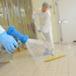 A close-up of a hand spraying water onto the white tile surface of a laboratory workplace while wearing blue latex gloves.