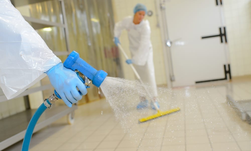 A close-up of a hand spraying water onto the white tile surface of a laboratory workplace while wearing blue latex gloves.