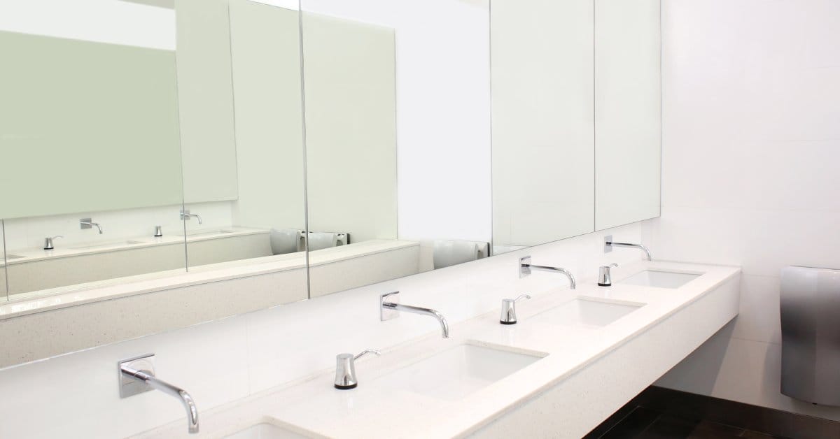A row of automatic faucets installed on the wall sit above a long line of white sinks in a public bathroom.