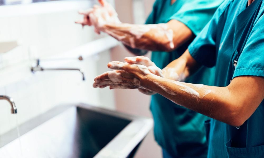 Two medical professionals wearing blue scrubs and washing their hands and forearms in a large silver sink.